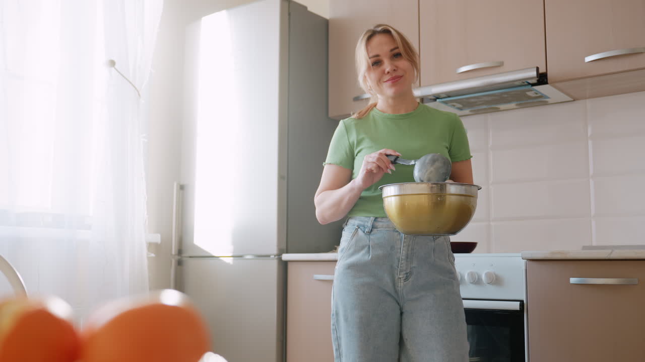 Donna con camicia verde e jeans che tiene una ciotola di metallo mentre mescola la farina con una frusta in una cucina luminosa, sorridendo mentre le arance si sfocano in primo piano vicino alla finestra con la luce del giorno che splende
