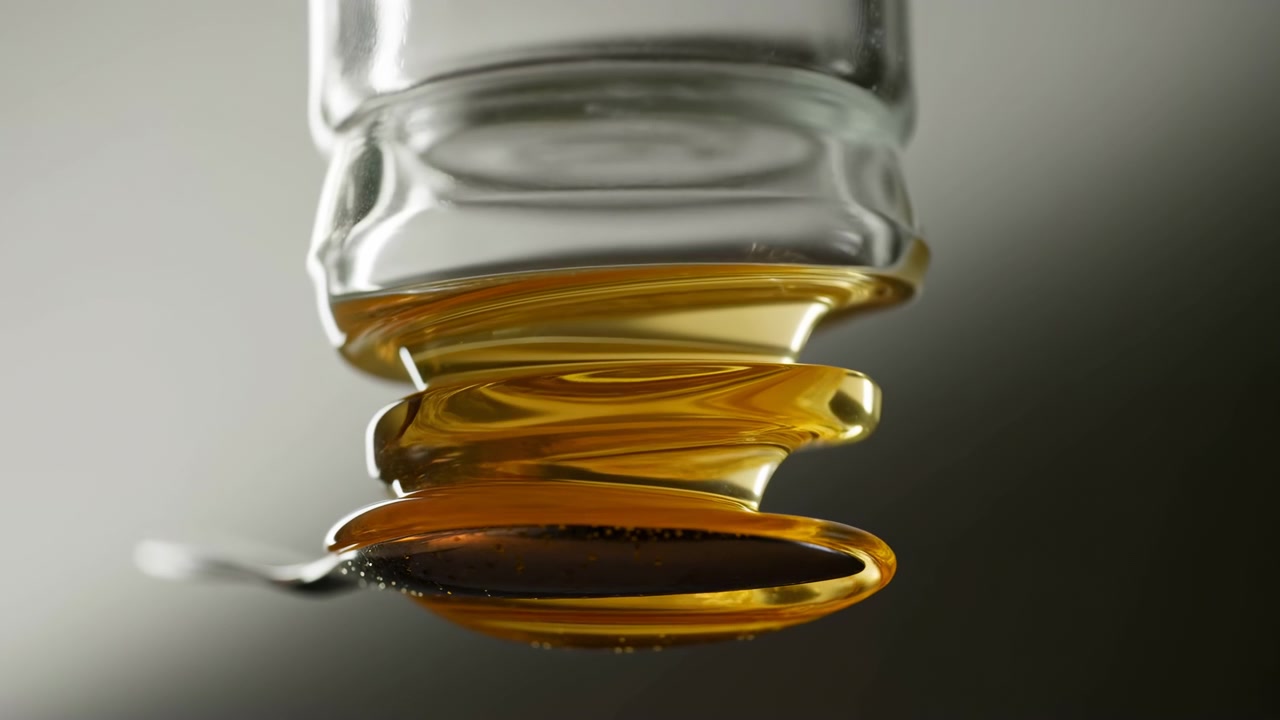 Close view of viscous amber syrup flowing from inverted clear glass jar into layered glossy droplets, showing thick strands, warm highlights, smooth reflections, and rich sticky texture