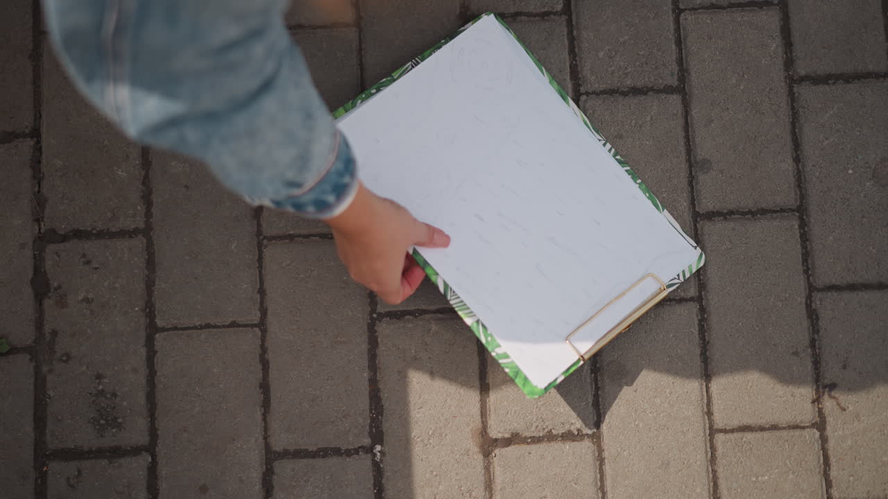 Mano che si allunga verso un portablocco sul marciapiede, manica di jeans e sneakers color verde acqua nell'inquadratura, luce solare che proietta l'ombra della mano sulla pagina bianca, movimento di raccolta in primo piano che suggerisce uno studente o un corriere che recupera appunti.