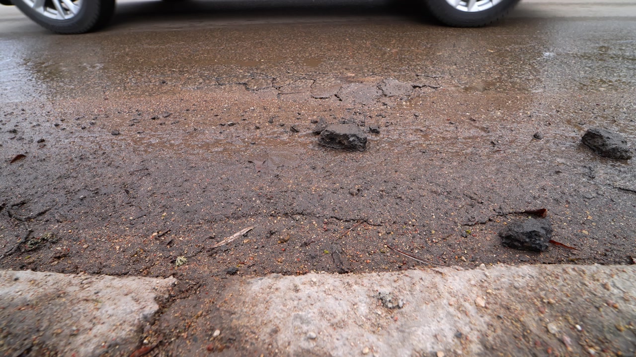 Close-up of dirty cracked asphalt with potholes, loose gravel, and winter road salt. Wet street surface shows mud and damage after snow and ice season.