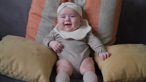 infant adorable baby girl sitting alone on sofa supported with cushions wearing a bib and headband in great mood smiling and lauphing