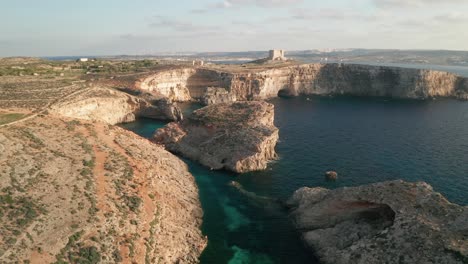 drone is flying over clear turquoise waters of malta's blue lagoon, unveiling rugged rock formations and coastal caves along como island's captivating shoreline