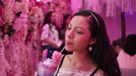 Hermosa-Mujer-Sonriendo-Y-Disfrutando-Bebiendo-Un-Colorido-Taro-Frappe-En-Una-Cafetería-De-Temática-Rosa-En-Tiempos-De-Pandemia-De-Coronavirus-Mientras-La-Gente-En-El-Fondo-Se-Toma-Selfies---Panorámica-Correcta