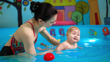 caucasian brunette woman is holding a baby in the swimming pool. little infant wants to cry but is calmed down by woman.