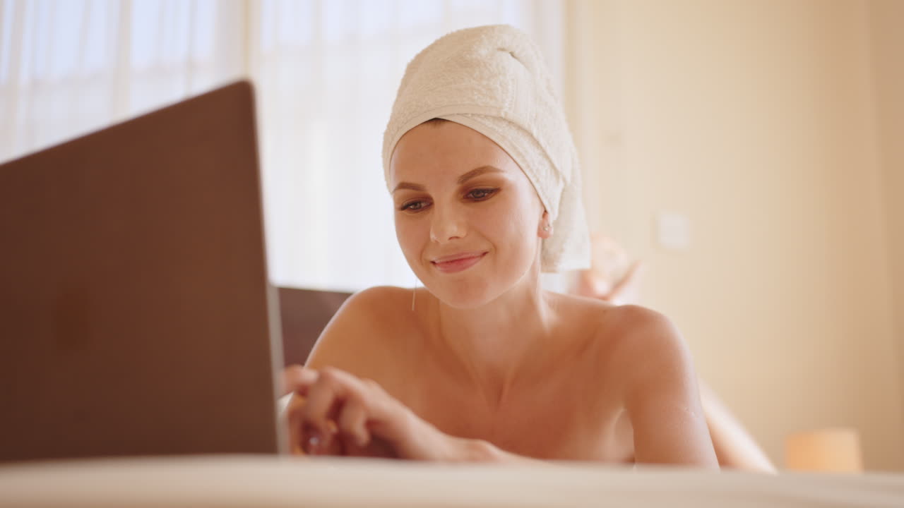 Splendida giovane donna avvolta in un asciugamano da bagno che digita al laptop mentre è sdraiata su un comodo letto