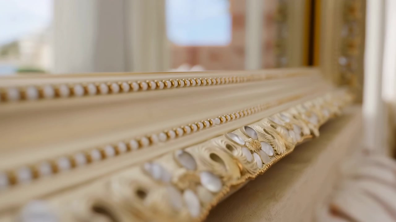 Primo piano estremo del bordo di uno specchio dorato e ornato che diventa più nitido mentre il primo piano rimane leggermente sfocato, rivelando perline intagliate, luce solare calda, eleganti dettagli interni e lo scenario del balcone riflesso.