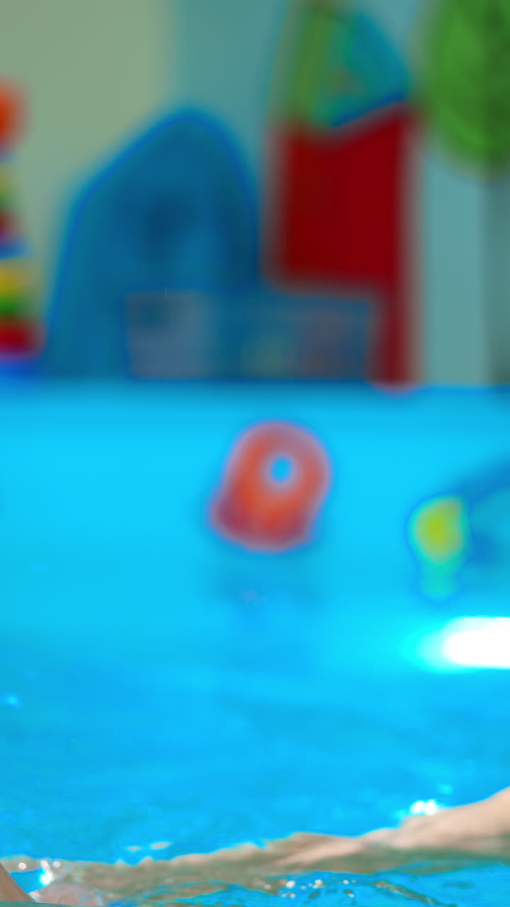 Smiling female coach holding a baby in the swimming pool. Blue-eyed newborn splashes water cheerfully. Vertical video