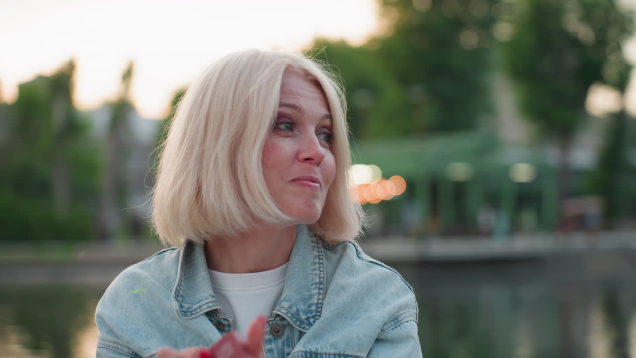 stylish woman outdoors by river at dusk cleans smudged paint from nose and applies eye makeup using compact mirror while wearing denim jacket and red nails reflecting gentle golden hour light
