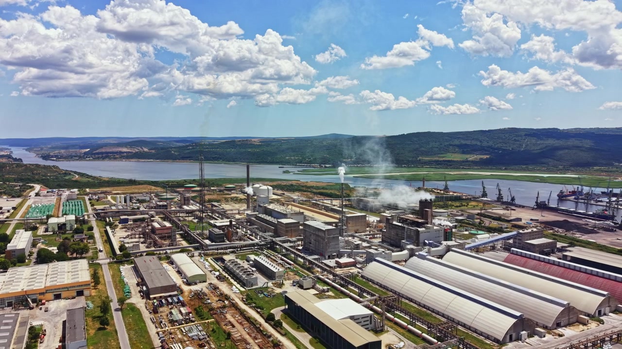 Sito industriale vicino a un fiume con edifici, macchinari e fumo durante una giornata limpida sotto un cielo blu con nuvole