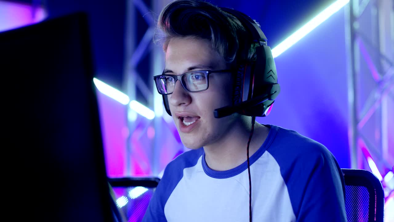 un joven adolescente jugador juega videojuegos en un torneo de deportes electrónicos o un café de internet. lleva gafas y auriculares y habla en el micrófono.