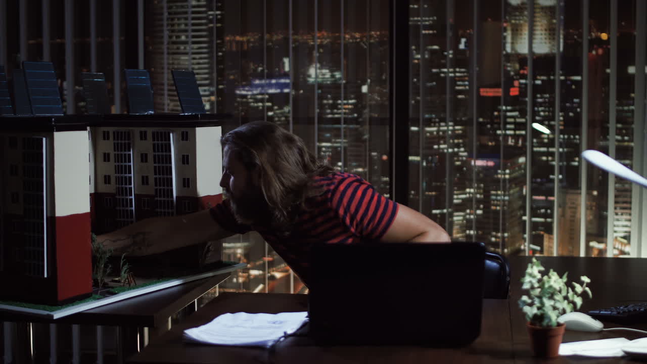Man working on a laptop and interacting with an architectural building model in a high-rise office at night