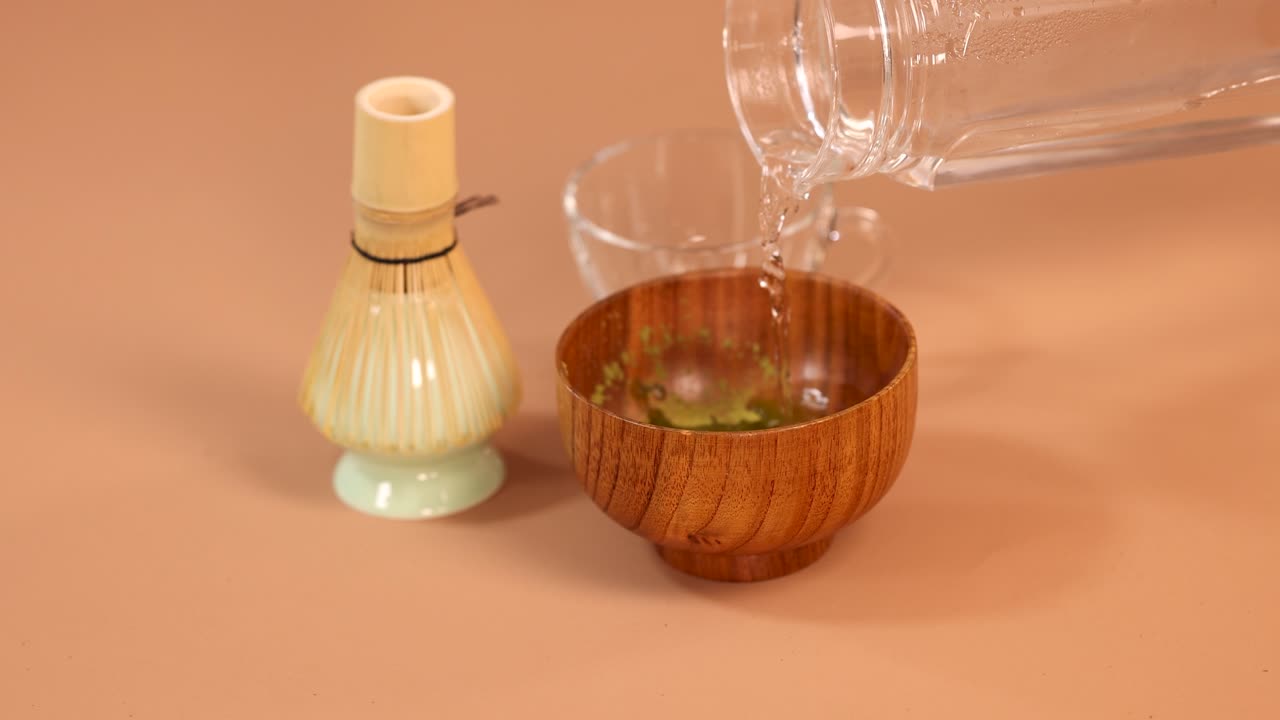 Hot water poured into a wooden bowl with matcha powder, traditional utensils in background