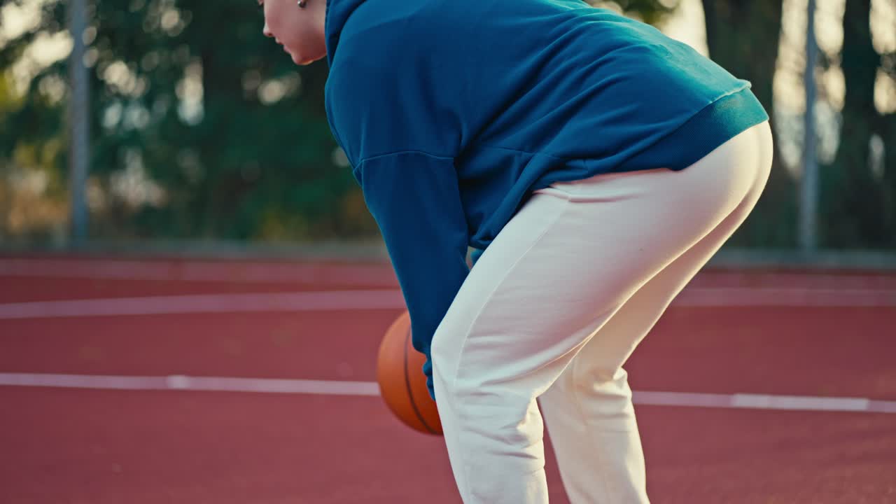 Close-up of a blonde girl in a blue hoodie and white pants hitting an orange basketball on the Red Street court in the morning during her workout