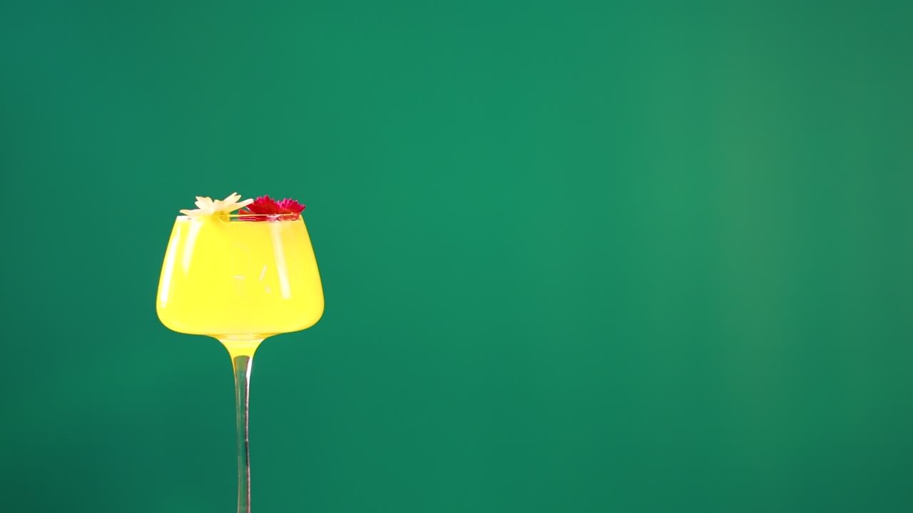 A vibrant yellow cocktail with floral garnish against a green screen backdrop. Static composition with bright, even lighting