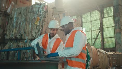 a brunette man with a beard in a white protective suit and an orange vest in a white helmet communicates with a colleague of a man with black skin who is working on a laptop near huge stacks of recycled paper at a waste recycling plant