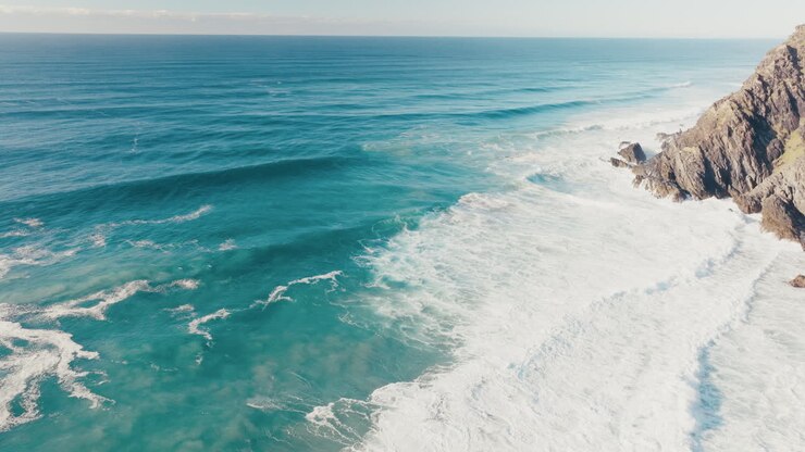 Onde turchesi dell'oceano che si infrangono verso un promontorio roccioso con schiuma bianca lungo la costa, ripresa aerea in tilt down.