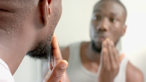 applying skincare, african american man looking in mirror and touching face at home spa