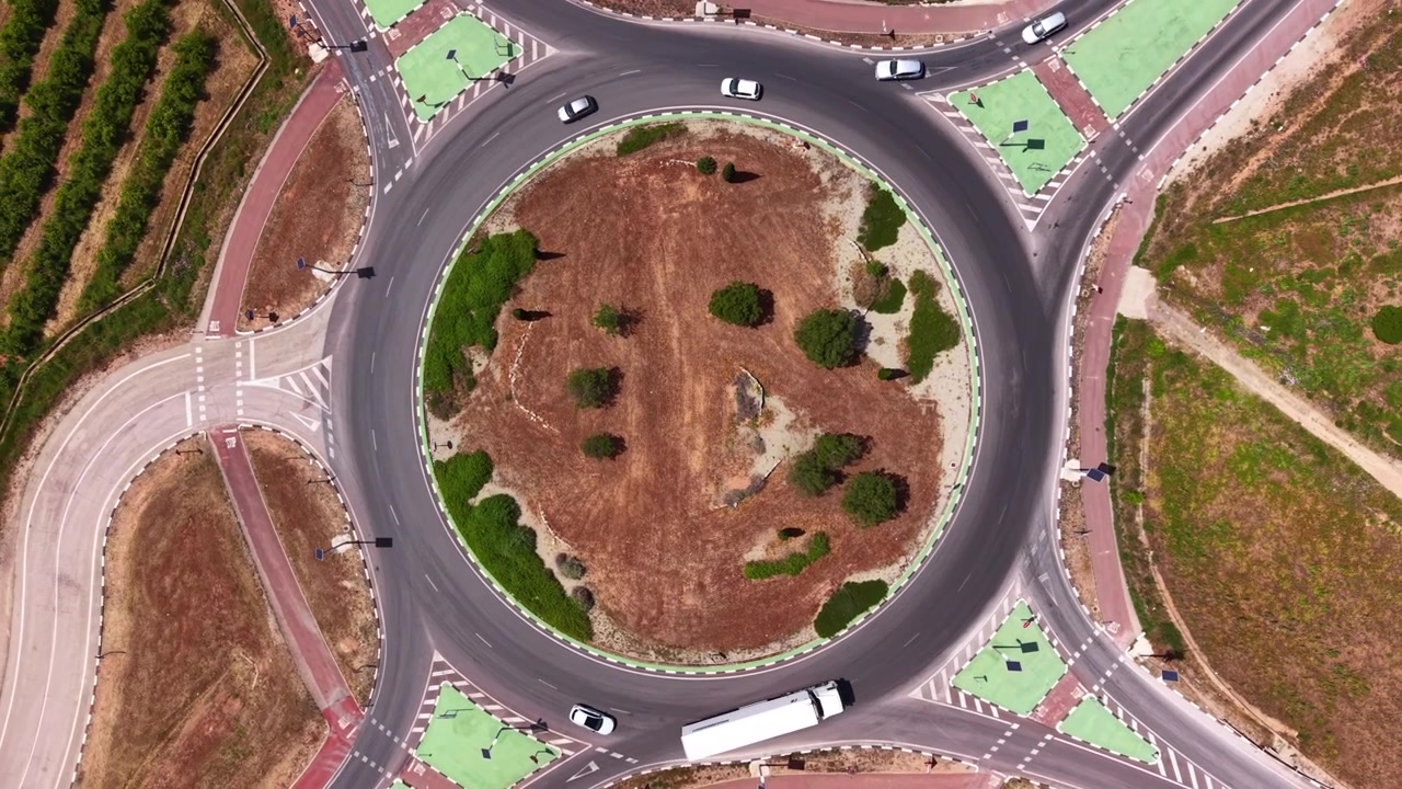 Top-down view of a large roundabout with circular landscaped island, vehicles including cars and truck navigating multiple entry roads with green pedestrian zones