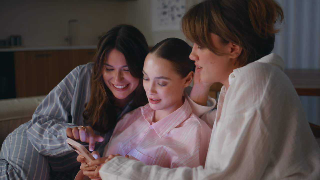 Girls friends enjoy messaging by mobile phone sitting apartment couch closeup
