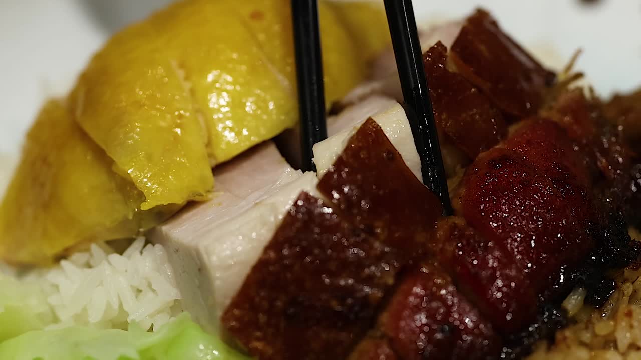 Close-up of chopsticks picking up succulent duck and chicken pieces over a bed of rice and greens.