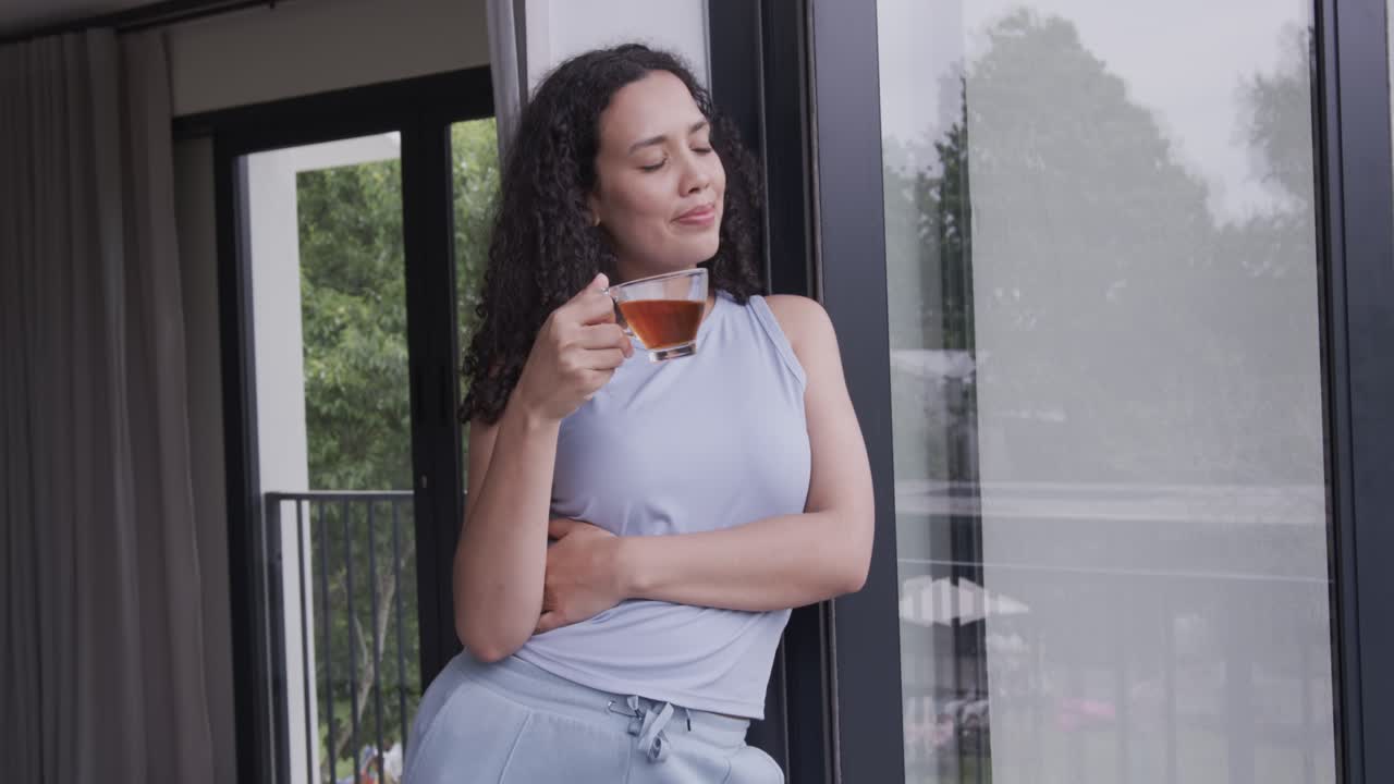 Donna afroamericana appoggiata a una porta scorrevole sul balcone, che tiene una tazza da tè di vetro e sorseggia alla luce nuvolosa.
