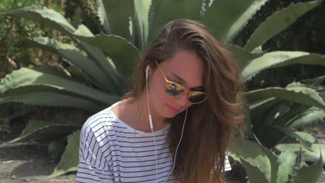 a young woman smiles and looks away from the camera. she is wearing sunglasses and has long blonde hair. she is standing in a garden with green plants and a white striped shirt.