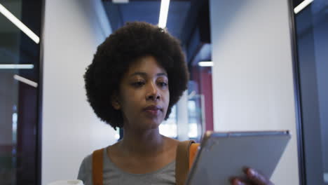 biracial businesswoman walking using digital tablet and drinking a cup of coffee in modern office