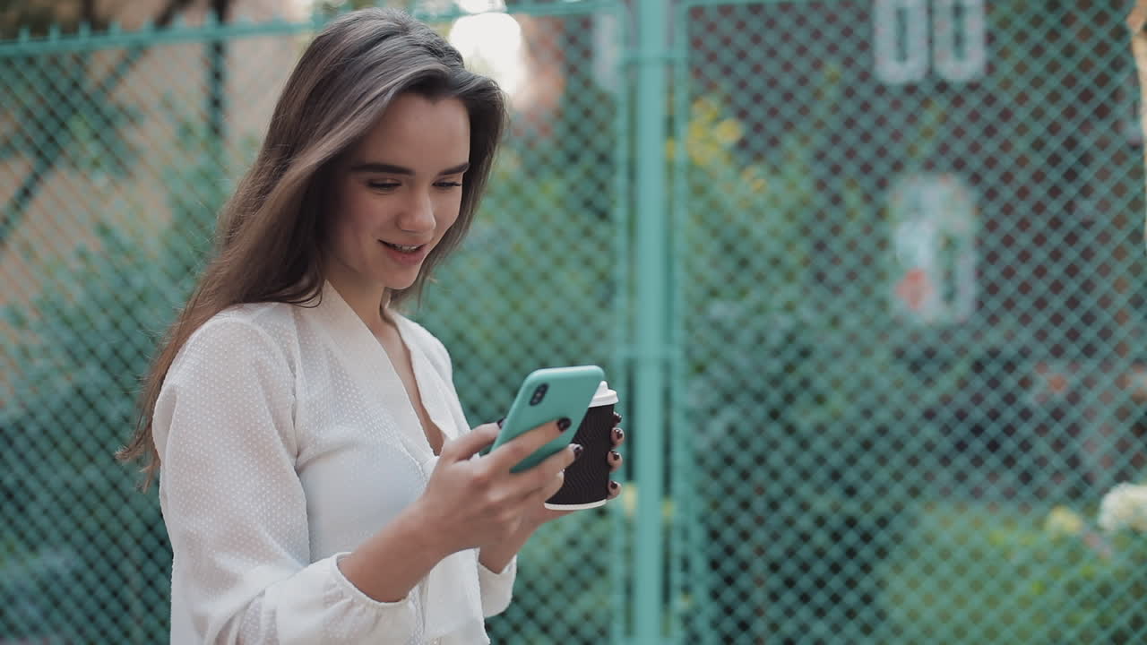 Woman using smartphone while holding a coffee cup near a fence