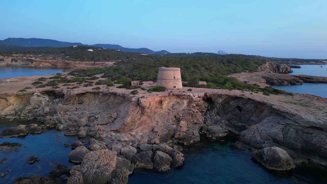 Donna in posa yoga che si gode il tramonto vicino alla Torre d'en Rovira, una torre di difesa costruita nel XVIII secolo a Cala Comte, Ibiza, Spagna. Spettacolare vista aerea con volo inverso del drone.