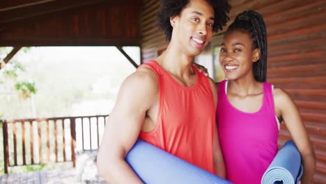 Portrait-of-happy-african-american-couple-holding-yoga-mats-in-log-cabin,-slow-motion