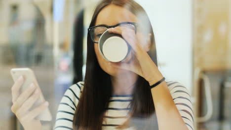 camera focuses on a brunette woman in glasses through the window drinking coffee and texting on the smartphone while she is sitting at a table in a cafe