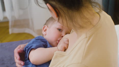 自宅のベッドに座って男の子に母乳を与える若い女性の肩越しのショット