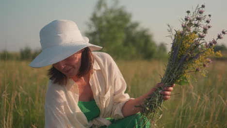 woman in white hat sitting in sunlit field gently admiring bouquet of wildflowers with peaceful smile, golden hour lighting enhances serene countryside atmosphere with soft focus on meadow background