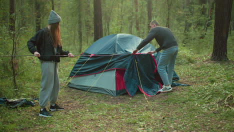 lady assists boy in forest as he inserts flexible pole into tent cover, both actively engaged in setting up camping shelter surrounded by trees and greenery, wearing casual outfits