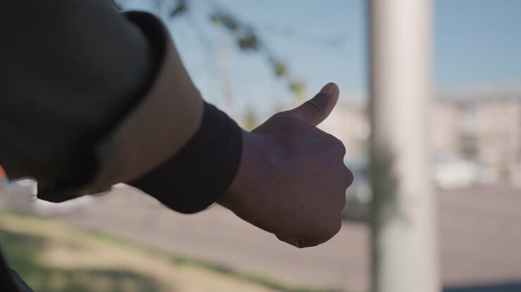 Close up of man giving thumbs up gesture from inside vehicle arm in dark jacket sleeve. Background shows blurred street scene with vehicles and buildings under daylight sky. Soft focus