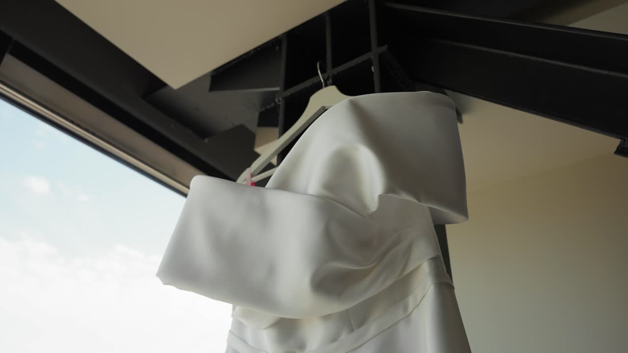 Close-up of an elegant white wedding dress hanging by a window in a modern loft, highlighting the luxurious fabric and minimalist architectural background