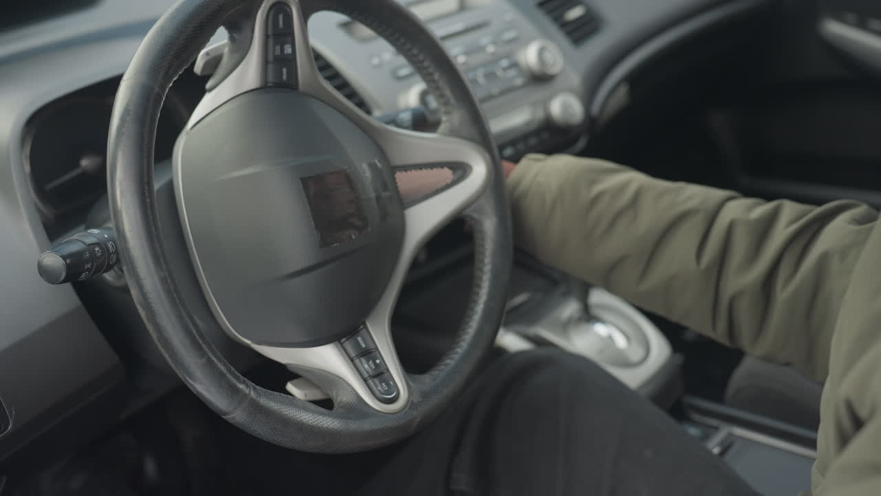 close up of person wearing jacket seated in driver seat holding key and inserting it into ignition to start vehicle, interior showing steering wheel, dashboard, and console controls in focus