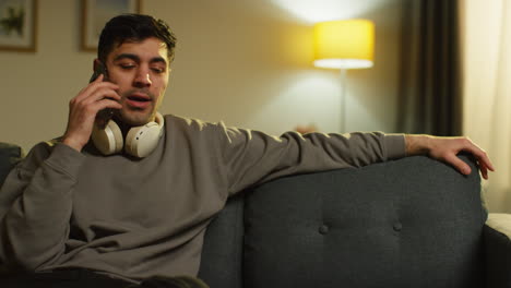 young man spending evening at home sitting on sofa with wireless headphones talking on mobile phone