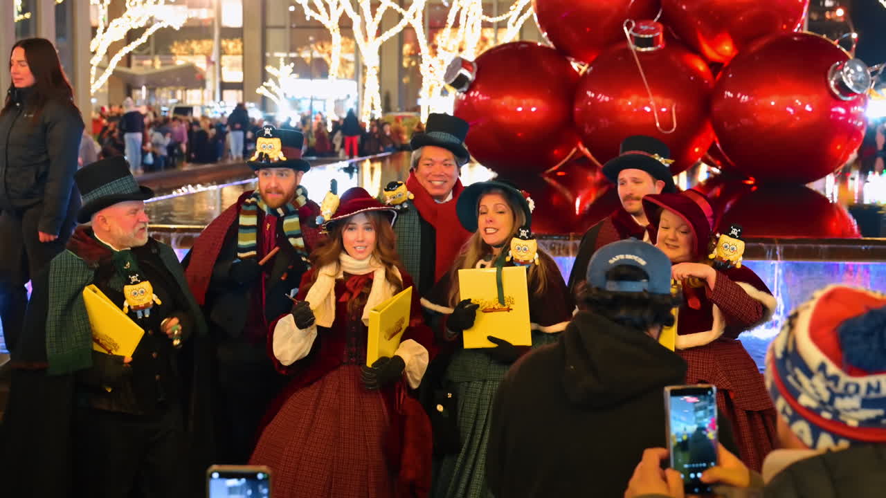 New York, USA, 5 december 2025: Viktorianska julsångare sjunger vid gigantiska röda julgransdekorationer på natten. En grupp julsångare klädda i viktorianska kläder sjunger bredvid en fontän, omgivna av gigantiska röda julgransdekorationer