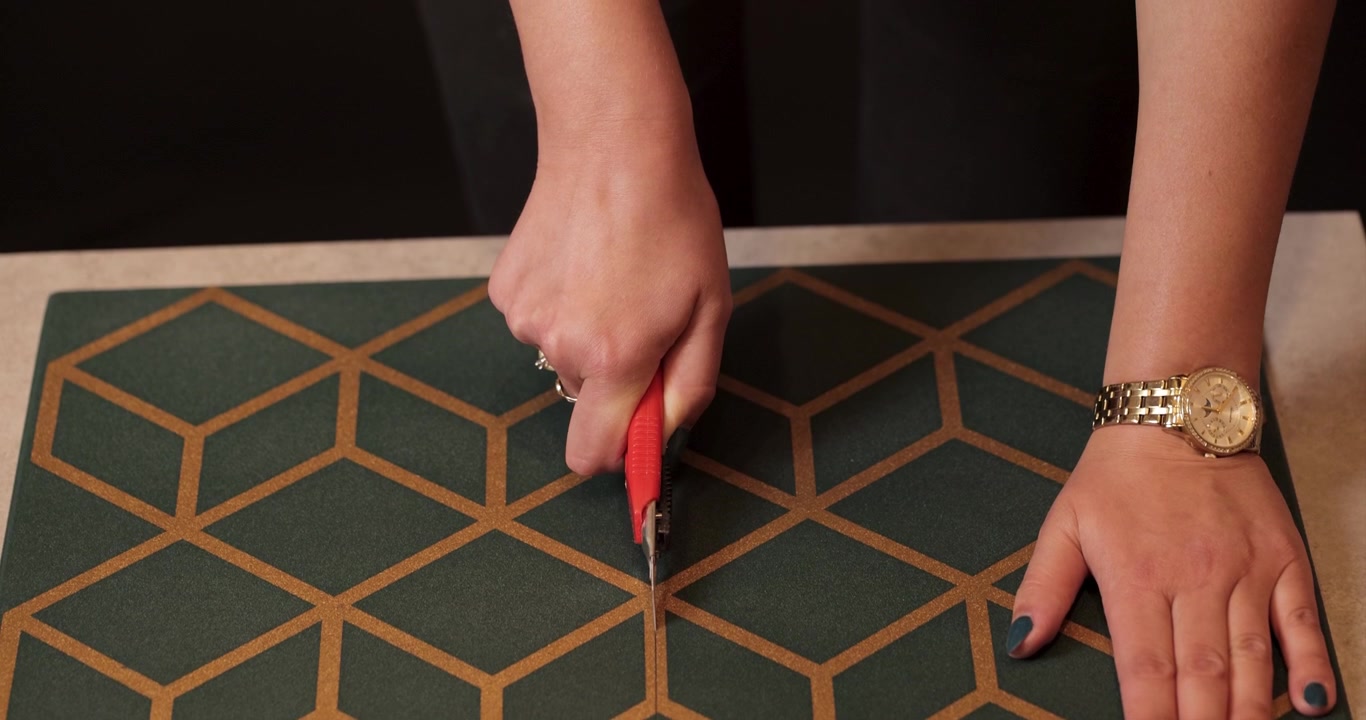 A close-up of the hands of a young Caucasian woman wearing a gold watch and rings, cutting a green and yellow styrofoam ceiling tile with a utility knife, shot in 4K resolution