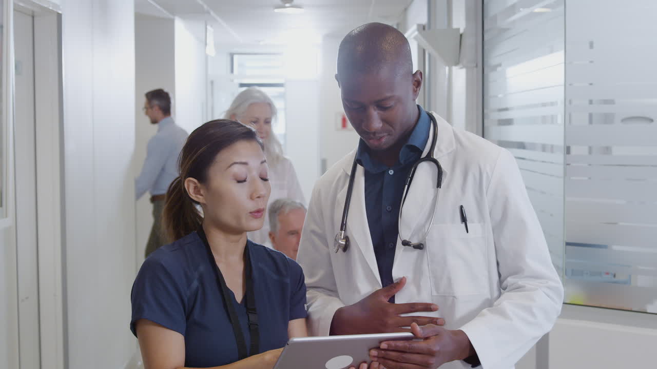 Doctor And Nurse Consulting Over Digital Tablet In Busy Hospital Corridor