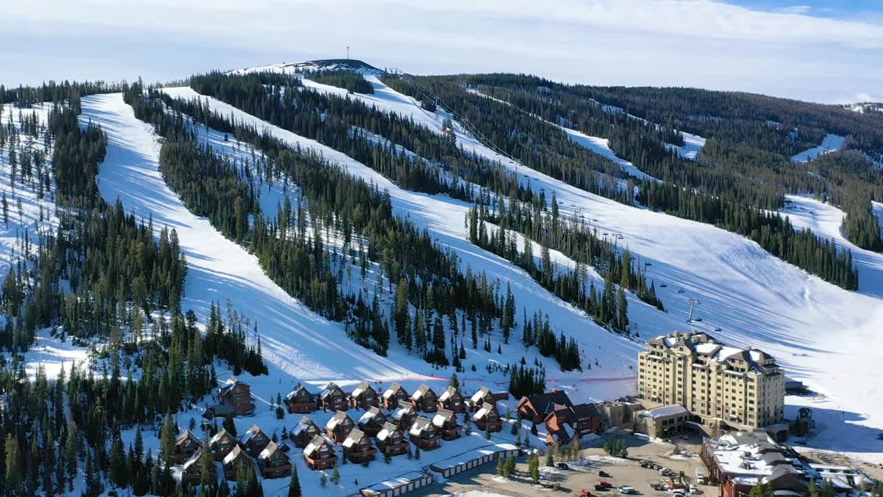 Flying above Big Sky, Montana, this aerial winter view highlights a ski resort surrounded by snowy peaks, pine forests, and nearby rental condos ideal for a relaxing cold-weather vacation