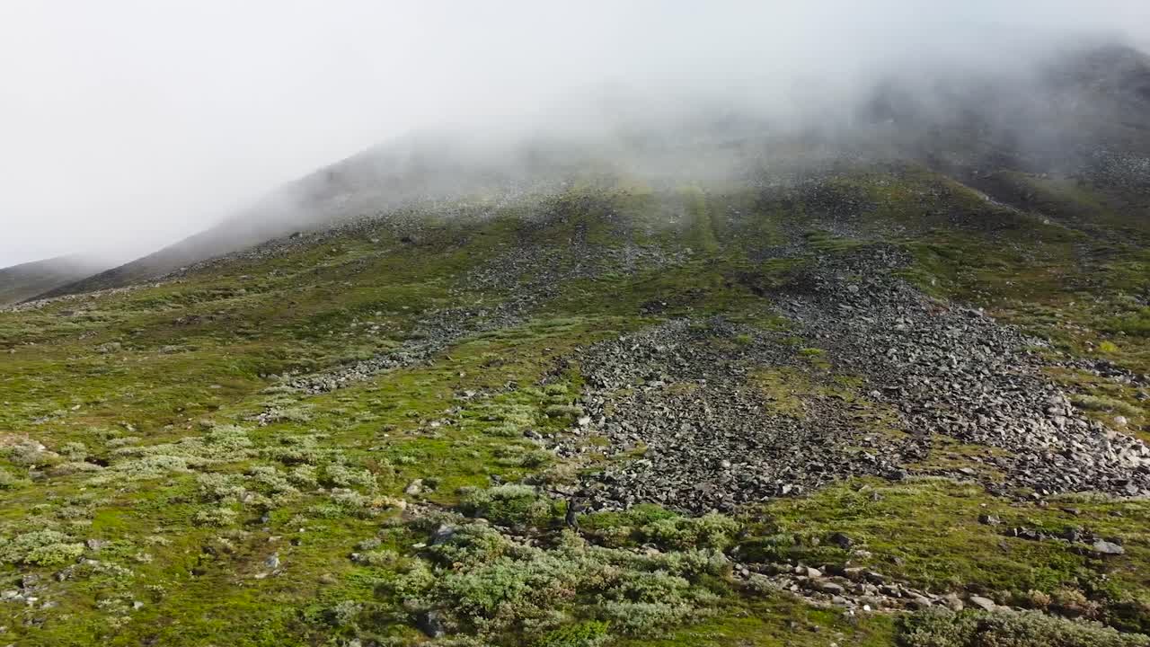 Aerial sideways movement footage of a person walking or hiking alone a rural and desolate path on a mountain side or hill slope that is grassy and rocky in Sweden during a sunny day, clouds visible.