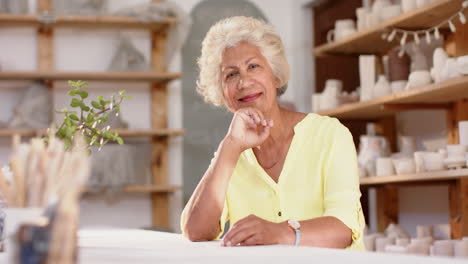 Feliz-Mujer-Birracial-Con-Cabello-Trenzado-Sentada-En-El-Escritorio-Y-Sonriendo-En-Un-Estudio-De-Cerámica,-Cámara-Lenta