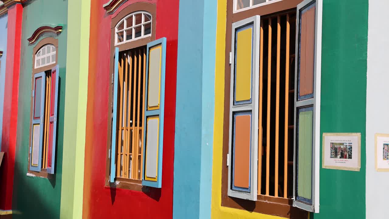 Camera pans along a vibrant, multi-colored shophouse exterior in bright daylight, revealing open windows and a lively urban street scene in Singapore