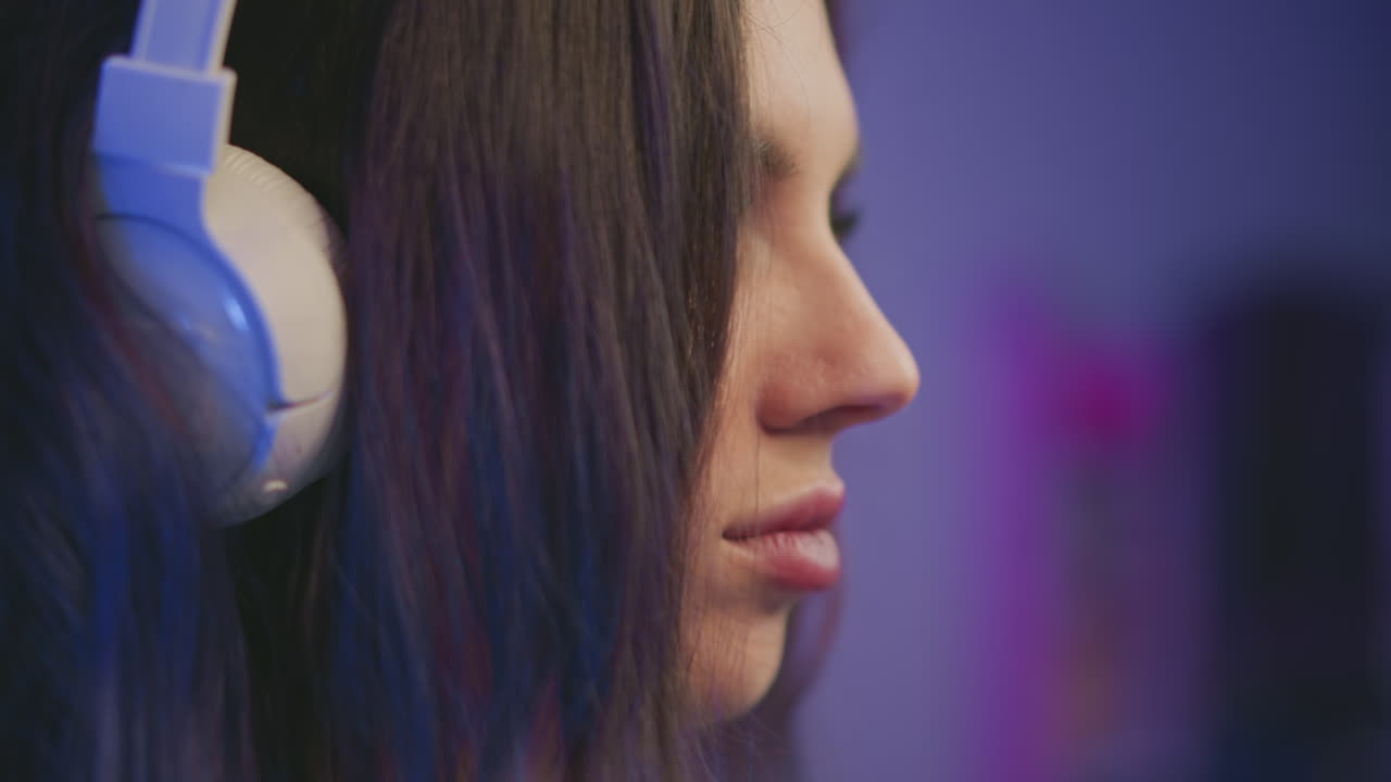 close up side view of young woman with long dark hair wearing white over-ear headset focusing on something off screen in softly lit room with purple ambiance and blurred background