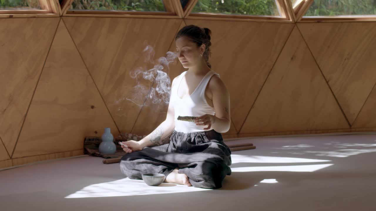 práctica de yoga en un estudio geométrico zen con una mujer latina abrazando movimientos tranquilos mientras quema un rollo de salvia para lograr la paz y la limpieza