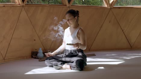 yoga practice in a zen geometric studio with a latina woman embracing calm movements as she burns a roll of sage to achieve peace and cleanliness