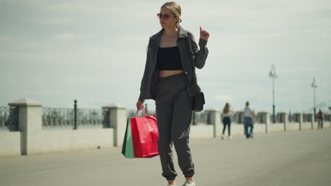 lady wearing black crop top under grey jacket dances, snapping her fingers while holding red, mint, and green shopping bags, with a hill on one side and blurred view of people walking behind her