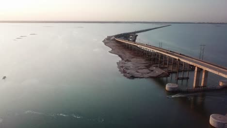 Vista-Aérea-De-Drones-De-Un-Pequeño-Barco-De-Pesca-De-Fondo-Plano-Que-Se-Desplaza-Bajo-La-Calzada-Conmemorativa-Jfk-Laguna-Madrea-Justo-Antes-Del-Atardecer